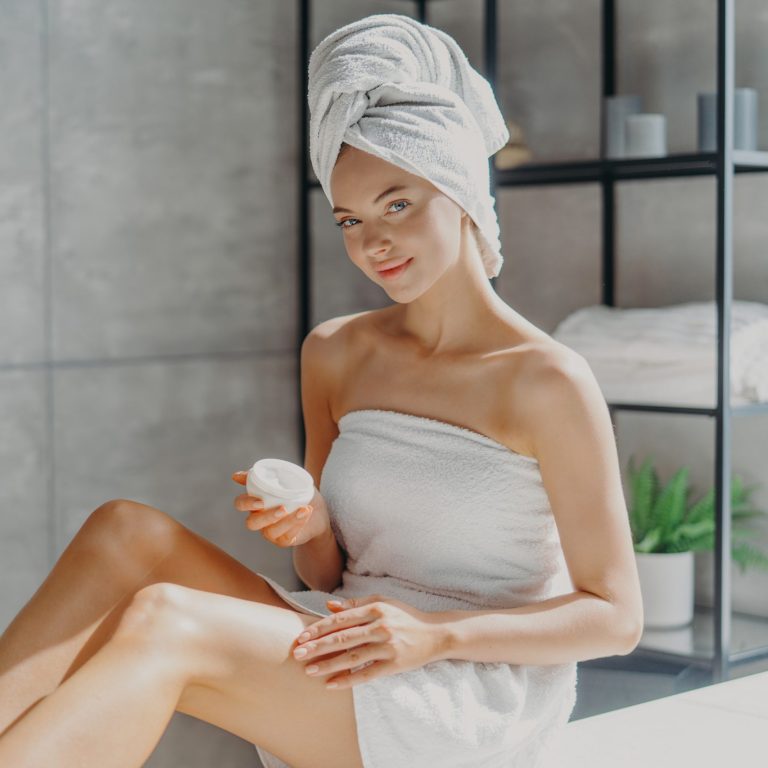 Horizontal shot of pretty woman applies cosmetic cream on skin, poses wrapped in towel, undergoes beauty procedures after taking shower, poses at home in bathroom. Daily hygiene and skin care concept lulur